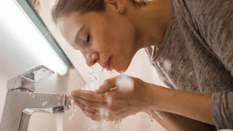 Getty Images Woman with her head next to a running tap splashes water over her face with her hands. 