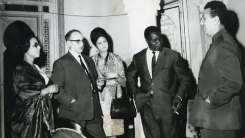 Eve Blouin Andrée, her husband, her daughter and the presidents talk in a group while wearing smart evening wear.