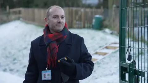 Hope Rhodes / BBC Head teacher Dan Crossman standing at the school gates. He is wearing a navy coat, checked scarf and there is snow on the grass behind him.