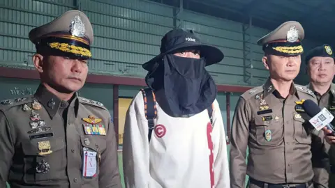 EPA-EFE Wang in a white sweater, wearing a black hat and a black face mask, flanked by two Thai police officials