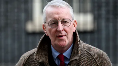 Reuters Northern Ireland Secretary Hilary Benn , he has short white hair, round-rimmed glasses and is wearing a brown herringbone-patterned overcoat over a blue shirt and red tie