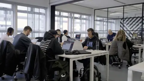 Staff at Hospital Records, sitting at grey desks in a warehouse-style office with their laptops. About seven members of staff are visible, young people wearing hoodies and T-shirts in mostly monochrome tones; some have chunky headphones on. At the far end is founder Chris Goss, an older man with grey hair, a Critall-style glass screen and a black-and-white abstract mural.