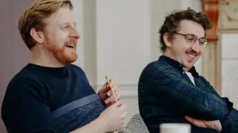 Michael Campbell Michael (left) in a navy jumper is holding a pencil and smiling and Oisín Kearney (right) who is in a shirt and is wearing glasses.