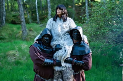 Claudia Winklemen being held up in the air by two assistants while filming The Traitors