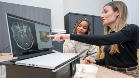 PA Media A woman with long fair hair and a black jumper points to a model of Ms Kaviya's skull on a computer screen. Ms Kaviya sits alongside her looking thoughtful.