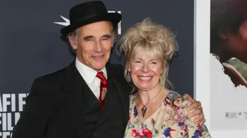 Getty Images Claire van Kampen, who has a blonde updo and wears a floral dress with large silver earrings and a pendant necklace, smiles as her husband Mark Rylance puts his arm around her shoulder at a film screening 