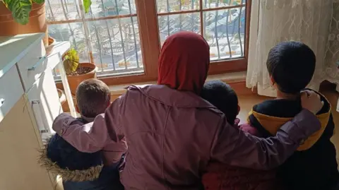 Niluper Niluper in a red headscarf and a mauve jacket with her arms around her three children, all of them in winter jackets - their backs are to the camera as they look out sit on the floor and look out the window.   