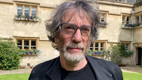 BBC Neil Gaiman pictured outside on the lawn of a property wearing a smart grey jacket and glasses