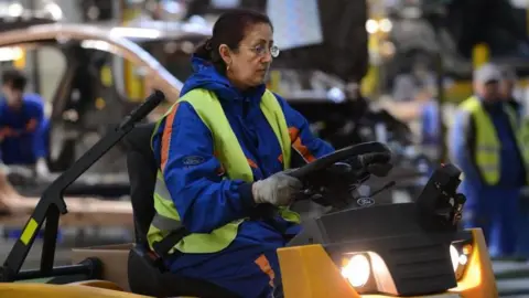 Getty Images A woman driving a vehicle inside  a factory, wearing a hi-vis vest on top of work overalls. 