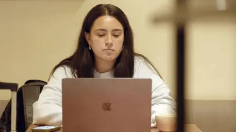 Maya, a woman with long brown hair, a nose ring and hoop earrings, wearing a light-coloured sweatshirt, sitting in the Hospital Records office in front of a MacBook with her iPhone to one side and a coffee cup on the other