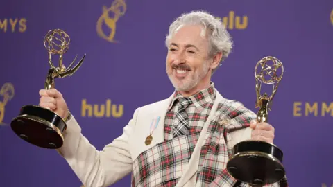 EPA Alan Cumming celebrates the Traitors winning two Emmys in 2024 - he is grinning broadly while holding two Emmy awards up, one in each hand. He is wearing a suit with a tartan plaid design.