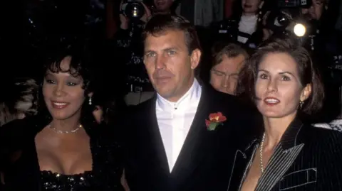 Getty Images Left to right: Whitney Houston, actor Kevin Costner and his wife Cindy Costner attending The Bodyguard Hollywood premiere in November 1992. They are all dressed in black  formal wear.