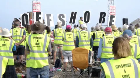 Protesters holding up signs that read 'Killing our communities'. They are wearing high-vis jackets with 