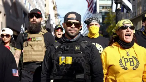 Getty Images Tarrio in sunglasses and a vest containing two yellow cans, flanked by others wearing Proud Boys gear