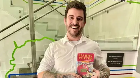 BBC Daniel Pearce standing in front of a glass panel holding the Harry Potter book with both hands. 