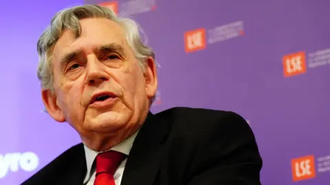 PA Media Gordon Brown speaking at a conference. He is wearing a suit and a red tie. An advertising board is visible behind him with the logos of several sponsors. 