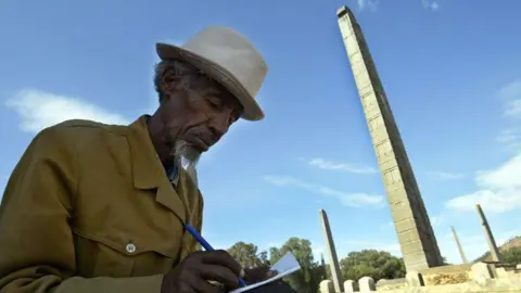 AFP A man in a hat with grey hair and and goatee bear writes in a notebook, behind him are the obelisks of Aksum