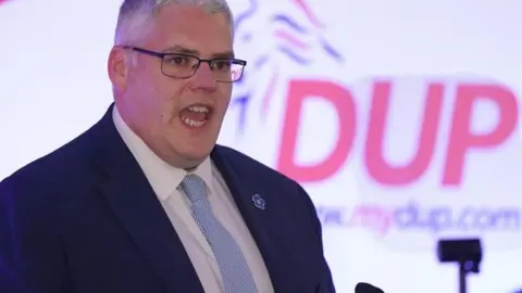 PA Media Gavin Robinson speaking at a DUP conference wearing a blue and white spotted tie, a white shirt and navy jacket. He is standing in front of a large, red-lettered DUP logo.