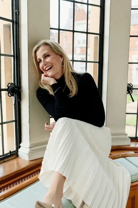 Christina Ebenezer/PA Media Sophie, who has long blonde hair and wears a black turtleneck with a pleated cream midi skirt, looks relaxed as she sits on a cushioned bench in a bay window and laughs while cupping her face with her hand 