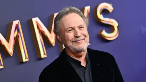 Getty Images Actor Billy Crystal smiles as he is photographed a red carpet event. He has grey hair and a heard and wears a dark suit.