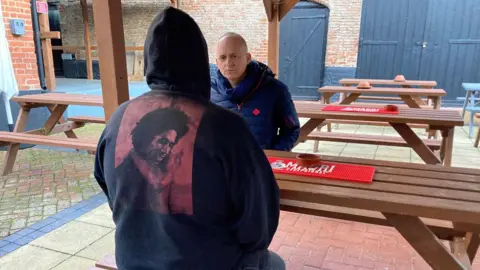 'Steve' is pictured from behind sitting on a picnic table in a paved pub garden. He is wearing a black hooded jumper, with the hood up, and a picture of Bob Marley printed on the back in red. Opposite him is Alastair, who is wearing a blue padded jacket and scarf.