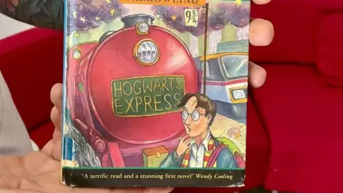 The front of the book which has a red train with the words 'Hogwarts Express' on the front. There is a illustration of a character, Harry Potter, stood in front of it with his hand to his mouth looking shocked. Along the bottom, there are the words 'A terrific read and a stunning first novel- Wendy Cooling'. 