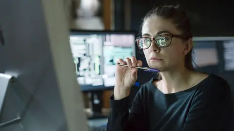 Getty Images Woman wearing glasses and black top, holding a biro under chin and looking at a computer monitor, with other screens seen behind her