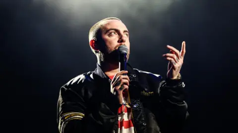 Getty Images Mac Miller on a dark stage with a spotlight overhead. He wears a black bomber jacket over a red and white striped top and holds a mic to his face with his right hand while pointing skywards with his left. 