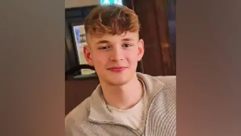 Hilary Rabbett George, a teenager with curly blond hair shaved at the sides and long at the top, smiles into the camera while wearing a beige coloured fleece open at the neck.