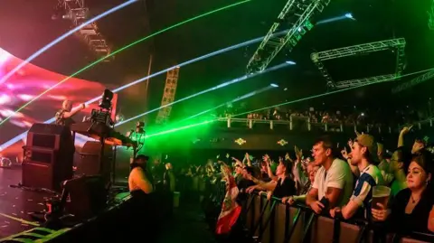 Getty Images People at a front row of a concert, right, watch Fatboy Slim on stage behind a DJ set of decks, left