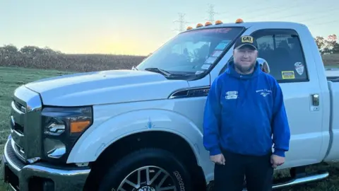 Ben Maurer Ben Maurer wears a blue hoodie and black CAT baseball cap in front of a white pickup truck