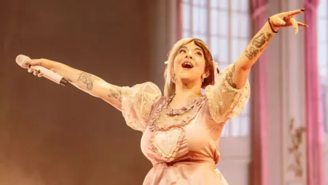 Getty Images Melanie Martinez perform in 2024 in Milan, Italy. She has her arms outstretched with her microphone held in her right hand and is looking up and smiling. She is wearing a pink dress, with short white puffed sleeves