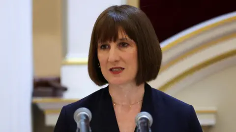 Reuters Rachel Reeves, a woman with a dark bob hair style, and a blue suit, stands in front of two microphones