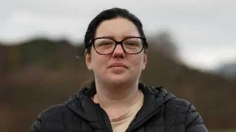 A photo of Tanya looking directly at the camera. It is a head and shoulders shot. She has dark hair tied back and glasses. She is wearing a big black coat. The photo is taken outdoors but the background is blurred. It looks like trees and a hill. 