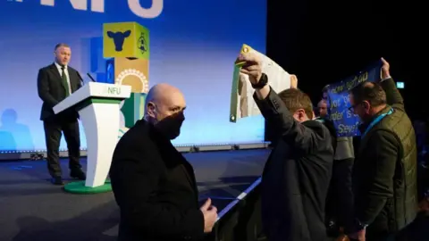 PA Media Side angle photo of protesters stood holding banners in front of Steve Reed, who can be seen in the background on the stage