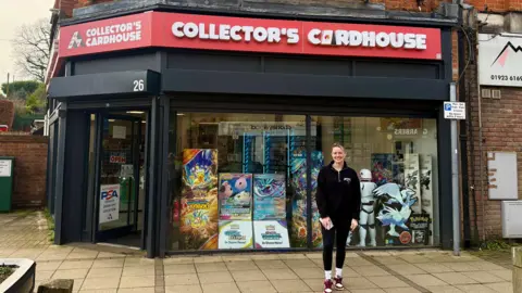 Danny Fullbrook/BBC A young blonde woman is smiling outside a shop which has a large red sign saying 