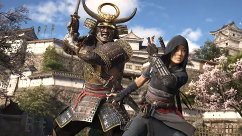 Ubisoft Screenshot shows two characters in the garden of a white Japanese castle as both adopt battle stances. One is a hulking warrior in bulky samurai armour and a horned golden helmet, holding a katana high and ready to strike. In front of him, a female ninja holds a throwing star in each hand, ready to launch them.