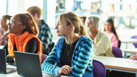 Getty Images Female caucasian student with blonde hair sitting in class in front of an open laptop at sixth form college listening and paying attention.