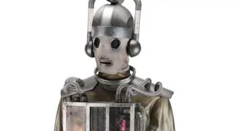 A close up of a Monda A Mondasian Cyberman which looks vaguely steampunk with a material like face. It has large silver headpiece and light-up box on its chest