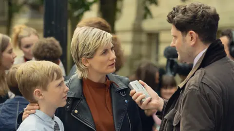 Netflix A still picture from a scene in the Toxic Town drama. Jodie Whittaker, in a black leather jacket and brown top, has her arm around a young boy. They are being interviewed by a male reporter in a brown coat who is holding a tape recorder near her mouth. They interview is taking place outside a court building.