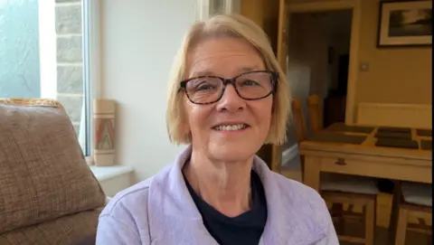 BBC A 72-year old woman with blonde hair and glasses is looking straight ahead and smiling.  She is wearing a lavender coloured jumper with a blue t-shirt underneath.  She is sitting in her conservatory, which backs onto the kitchen.