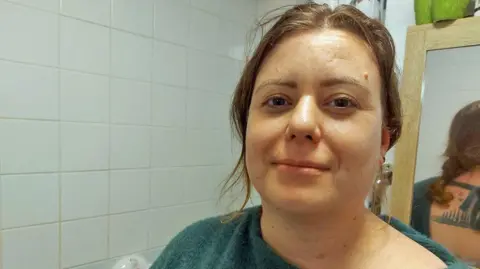 BBC Headshot of Sam in her bathroom. She has wavy brown hair tied back, with some strands framing her face. She is wearing a green off-the shoulder jumper - and reflected in the mirror behind her you can see it is tied at the back and she has a tattoo. The wall behind her has white tiles, there's a shower curtain and wooden framed bathroom cabinet. 