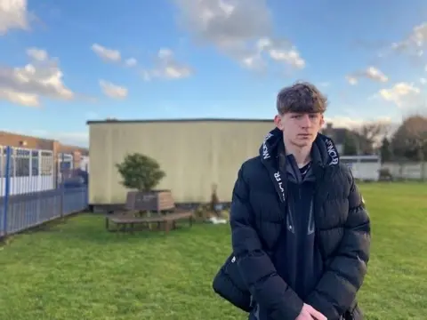 Lincoln O'Neill stands on the grass in school grounds. He is dressed in a black winter puffer jacket. There are buildings in the background.