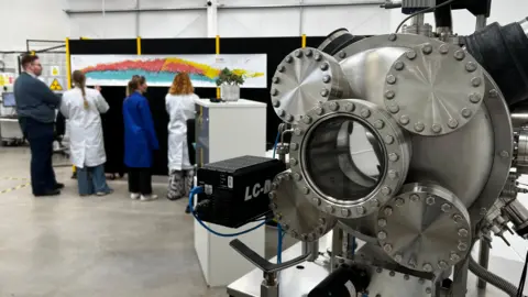 BBC Four research scientists stand behind a shiny steel scientific device. It has five discs, the central one of which has a glass window. The scientists, two in white lab coats, stand behind it looking at a colourful periodic table stuck on a wall.