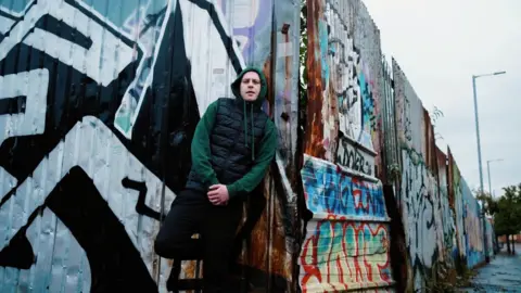 Skeet89 Young Spencer, wearing a green hoodie with a black, puffer gilet. He is leaning against a corrugated metal fence with graffiti on it.