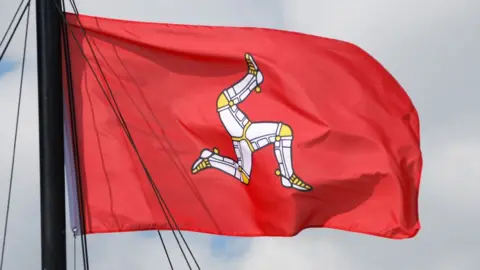 Getty Images The Isle of Man flag, coloured red with the triskelion in the middle, flies in the wind. The triskelion is an image of three legs in armour, fitted with golden spurs.