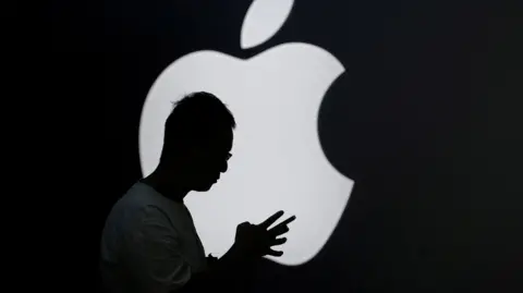 Reuters Silhouette of a man holding a smartphone in front of a lit-up Apple logo.