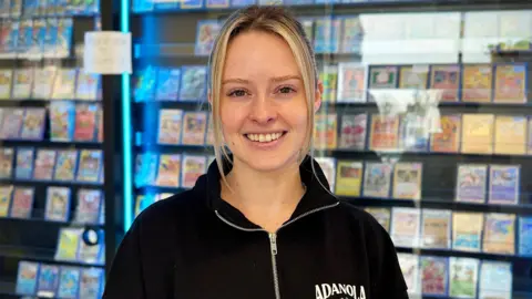 Danny Fullbrook/BBC A young blonde woman is smiling, behind her is a wall covered in Pokemon cards behind a class case.