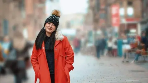 MKC Photocreations A photo of Lisa looking directly at the camera as she stands outside on a pedestrian street. The background is blurred out and Lisa's full length red coat stands out from the background. She has a black bobble hat on her head and has long dark hair. She is smiling at the camera.