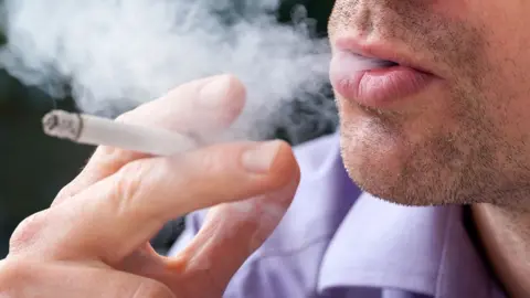 Getty Images Close-up of a man smoking a cigarette. He has a cigarette in his hand and is blowing smoke out of his mouth.